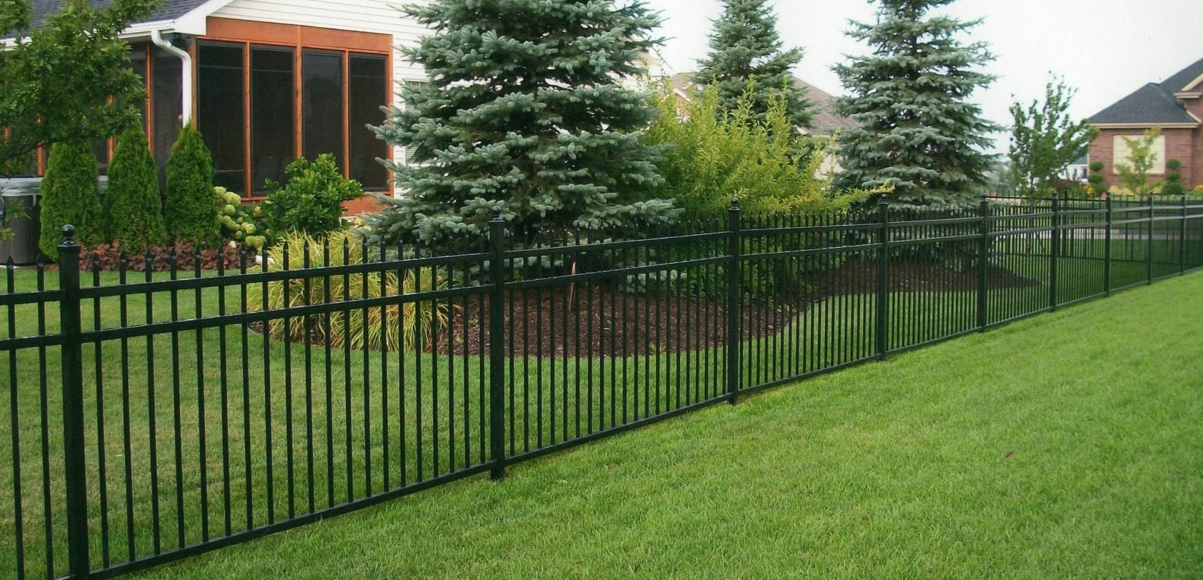 Black metal fence surrounding a grassy yard with evergreen trees. A house is in the background.