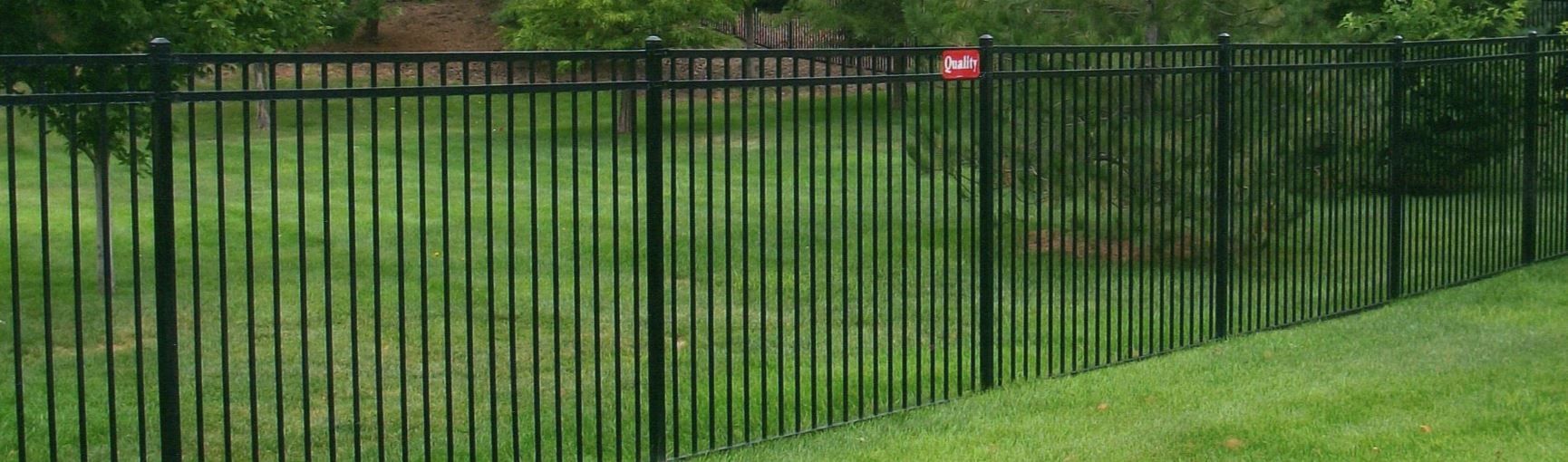 Black metal fence on a grassy hill with trees in the background. A red sign is attached to the fence.