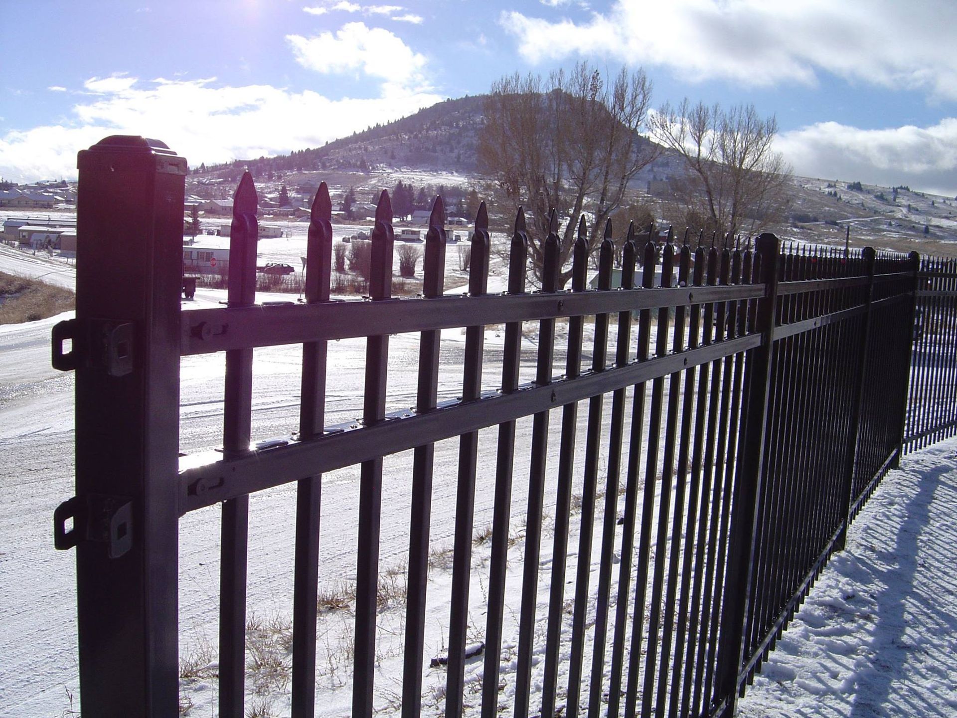 Black metal fence with pointed tops in snowy landscape, mountain in the background under a blue sky.