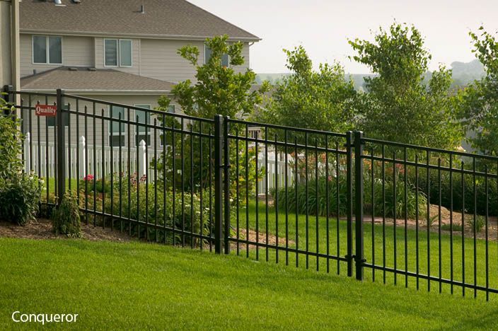 Black metal fence on a grassy hill, with a house and trees in the background.