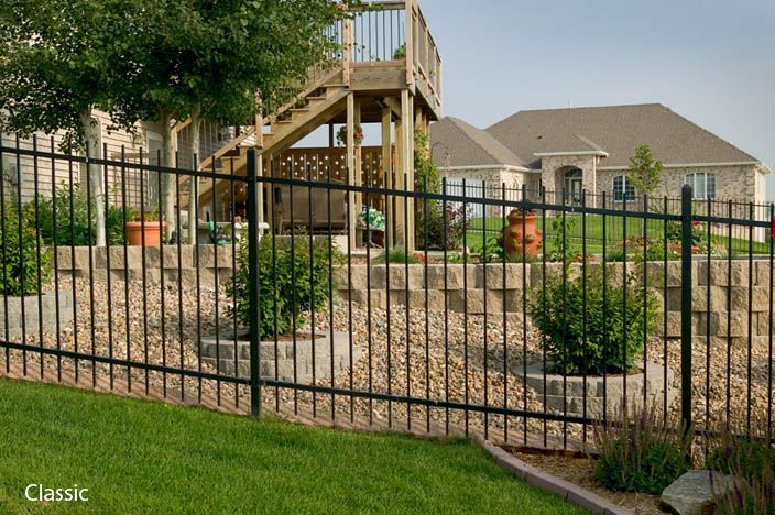 Black metal fence in front of a house with a wooden deck and a stone retaining wall with landscaping.