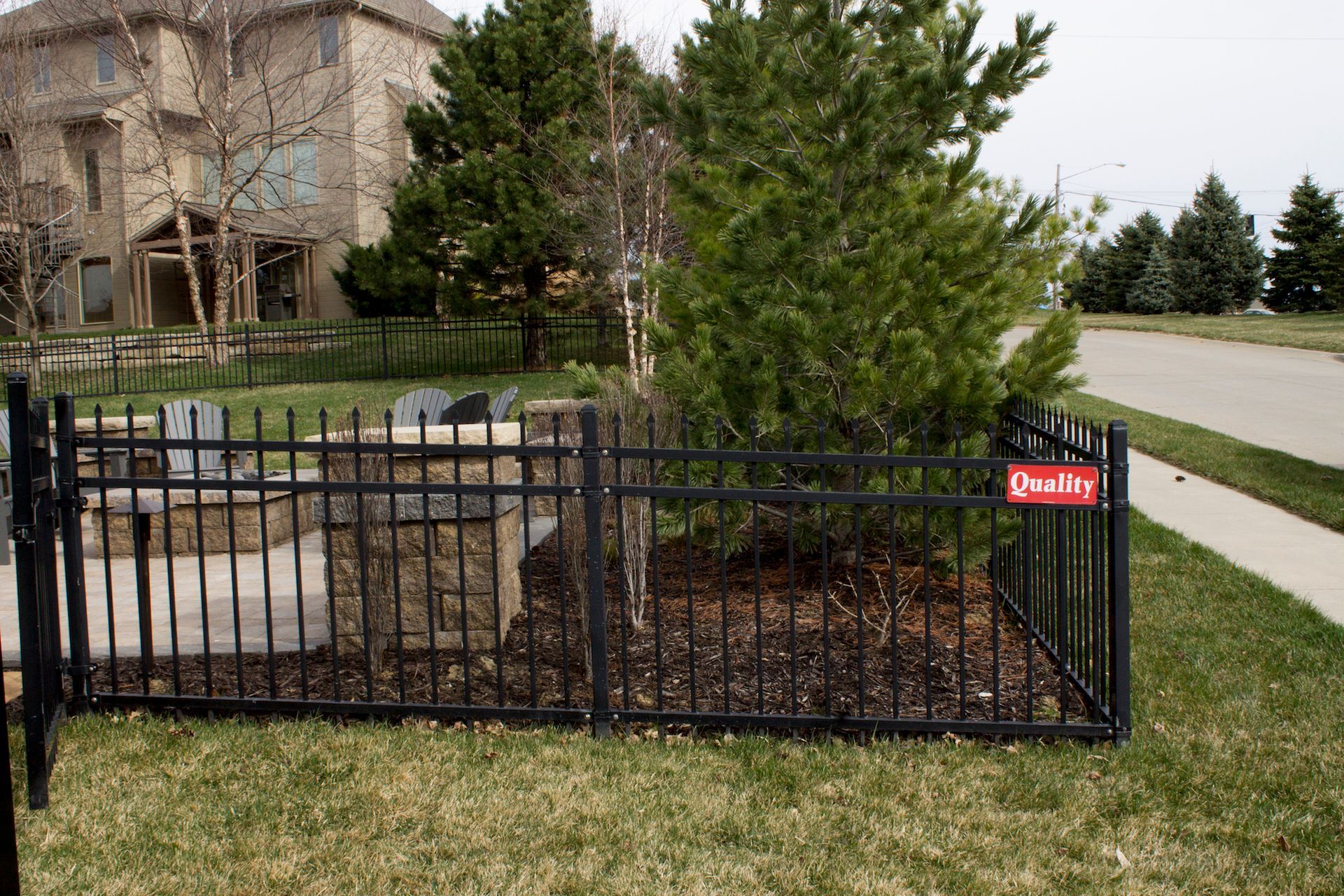 Black fence encloses a small yard area with a tree and a stone structure; grass and pavement in view.