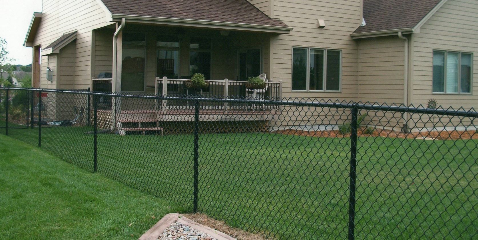 Black chain-link fence surrounds a grassy backyard, adjacent to a beige house with a porch.