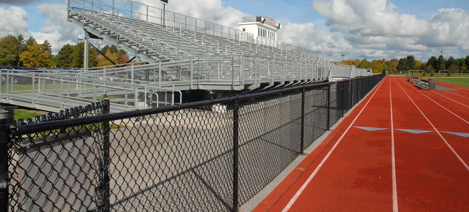 A track and field stadium with a red running track, bleachers, and a chain-link fence on a sunny day.