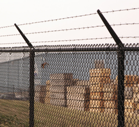 Black chain-link fence with barbed wire on top, guarding a building with stacked brown materials.