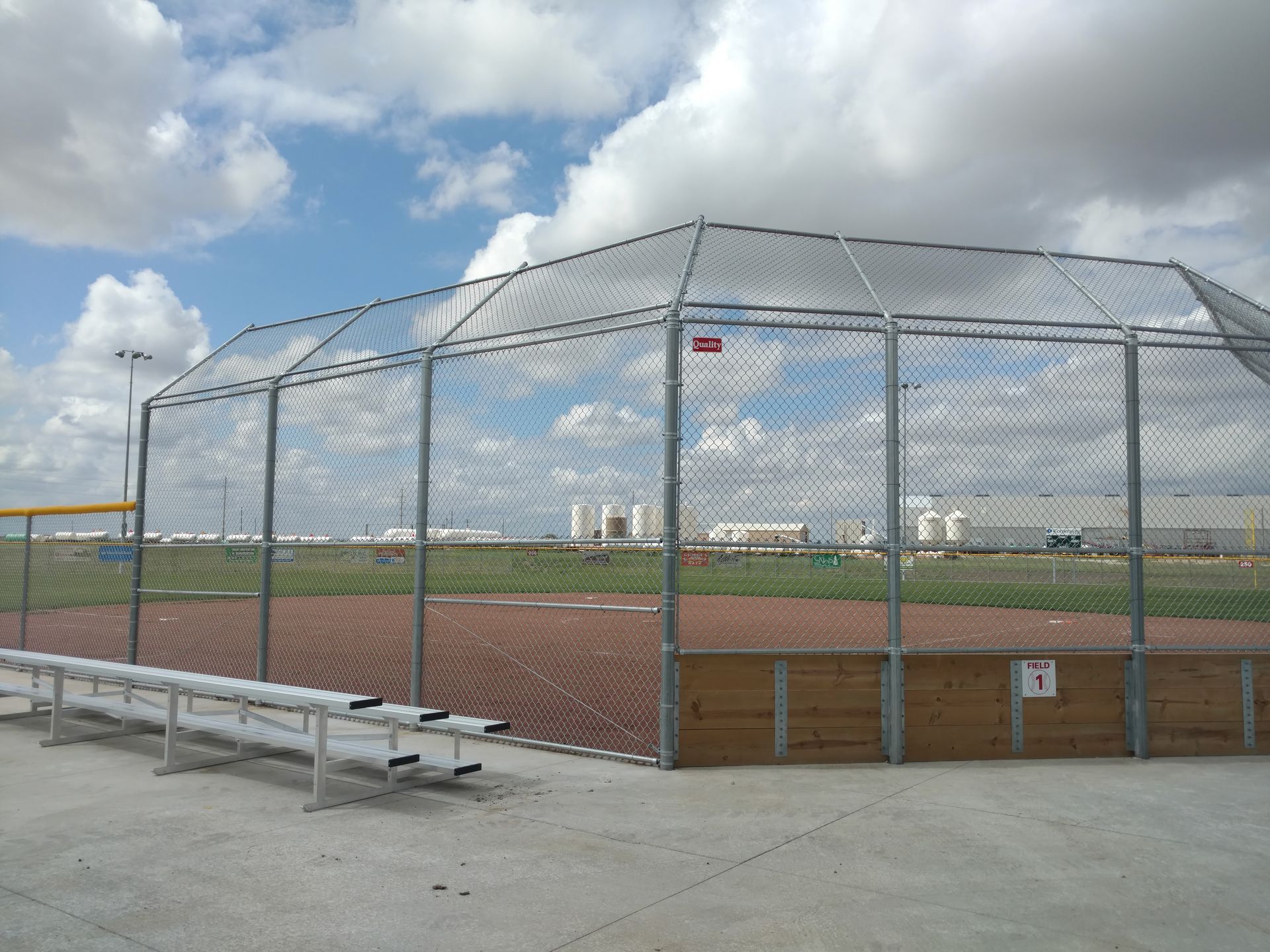 Baseball batting cage with chain-link fencing, red clay infield, and metal bench seating under a cloudy sky.