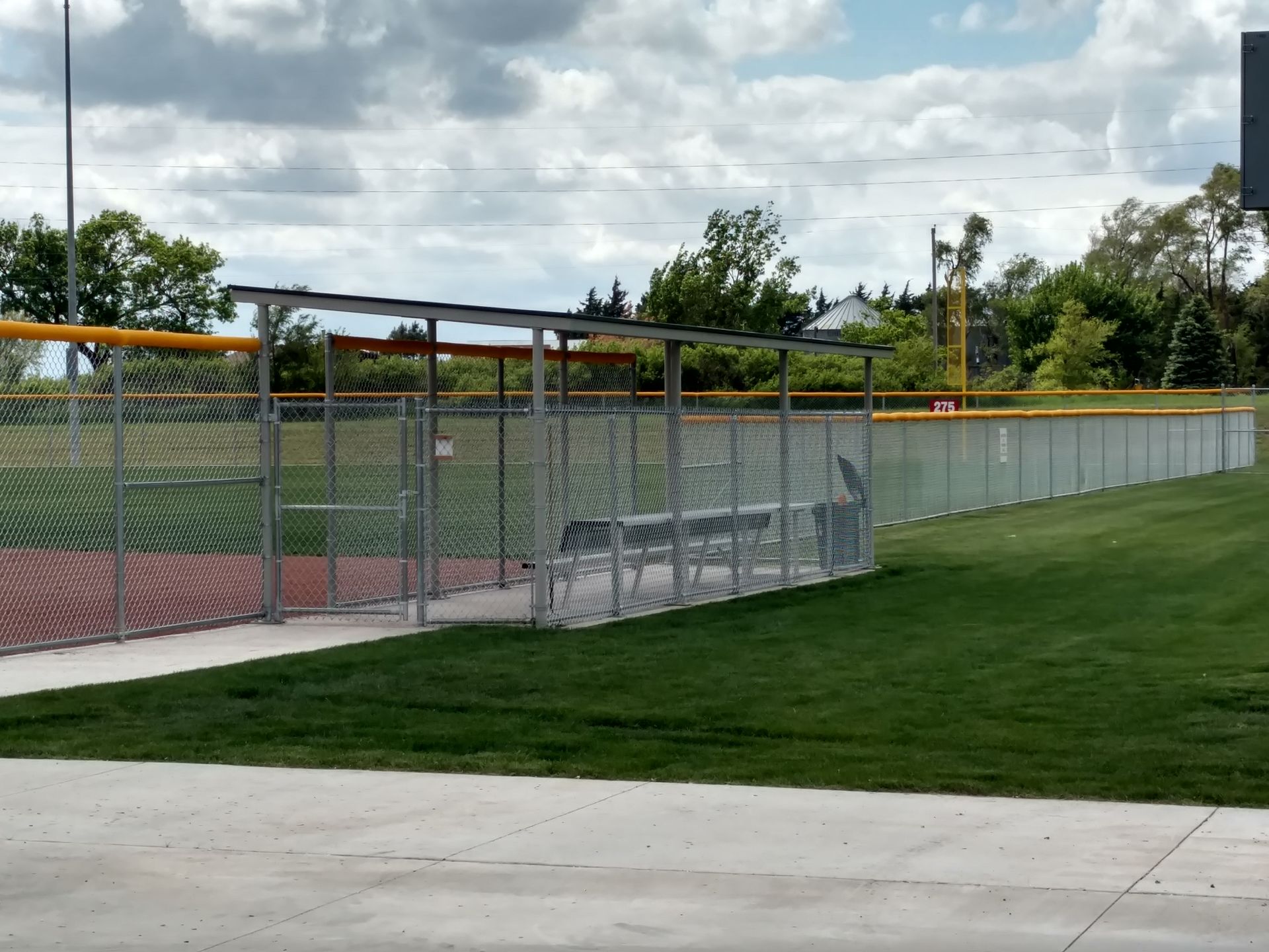 Baseball dugout behind a chain-link fence on a green field under a cloudy sky.