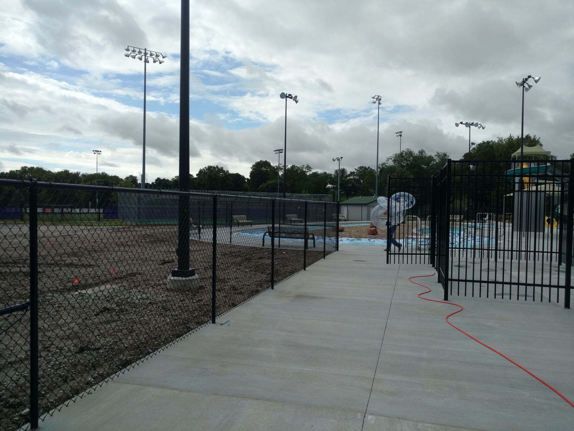 Fenced pathway leads to a water park area, with pools, a slide, and field lights, under a cloudy sky.
