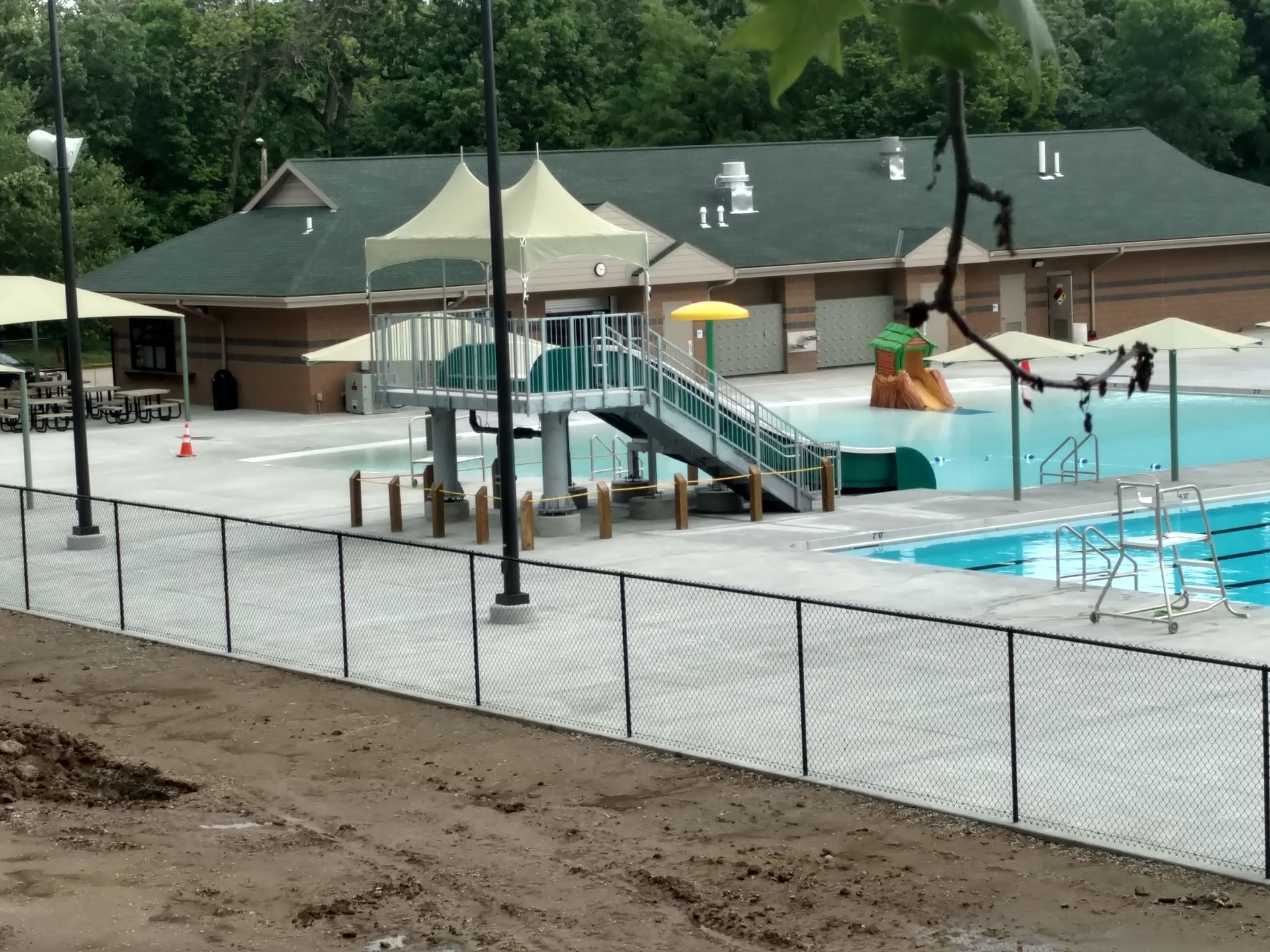 Pool area with slides, fence, and building in the background. Blue water and a light-colored patio surround the pool.