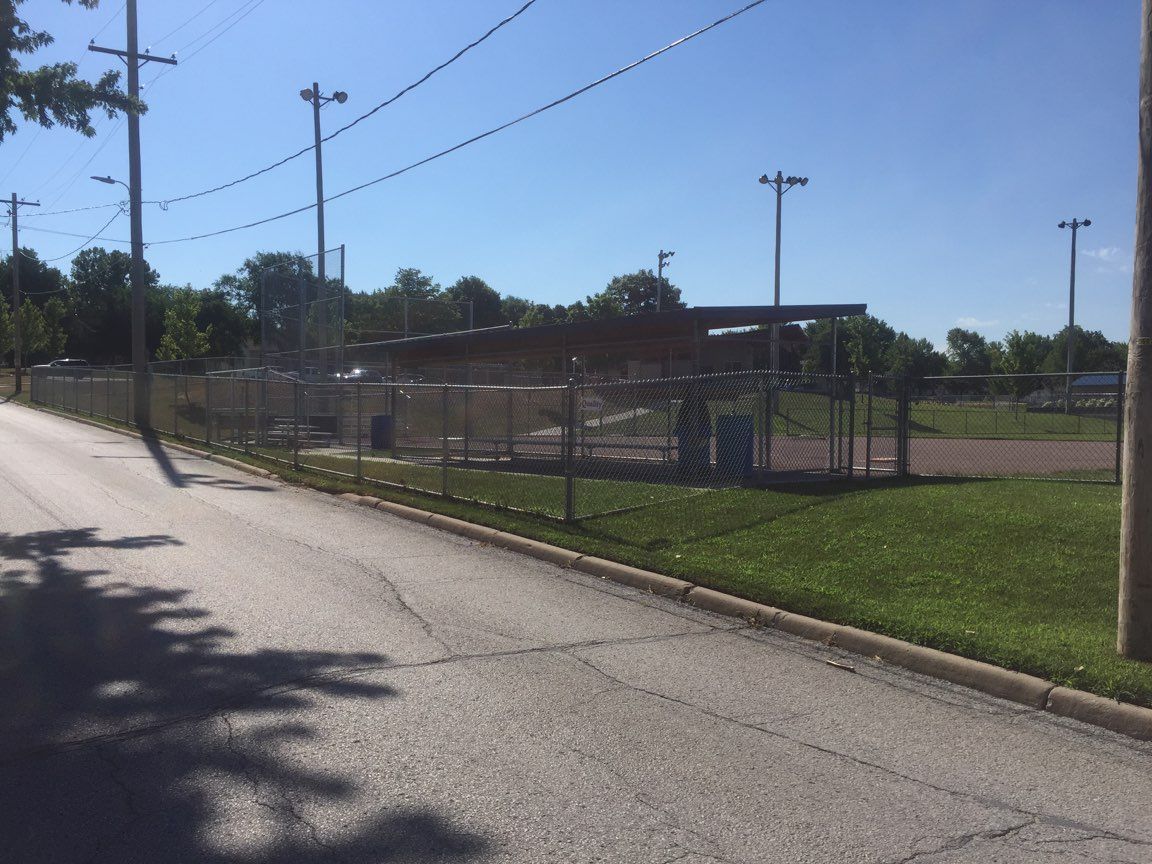 Baseball field with chain-link fence, dugout, and overhead lights on a sunny day.