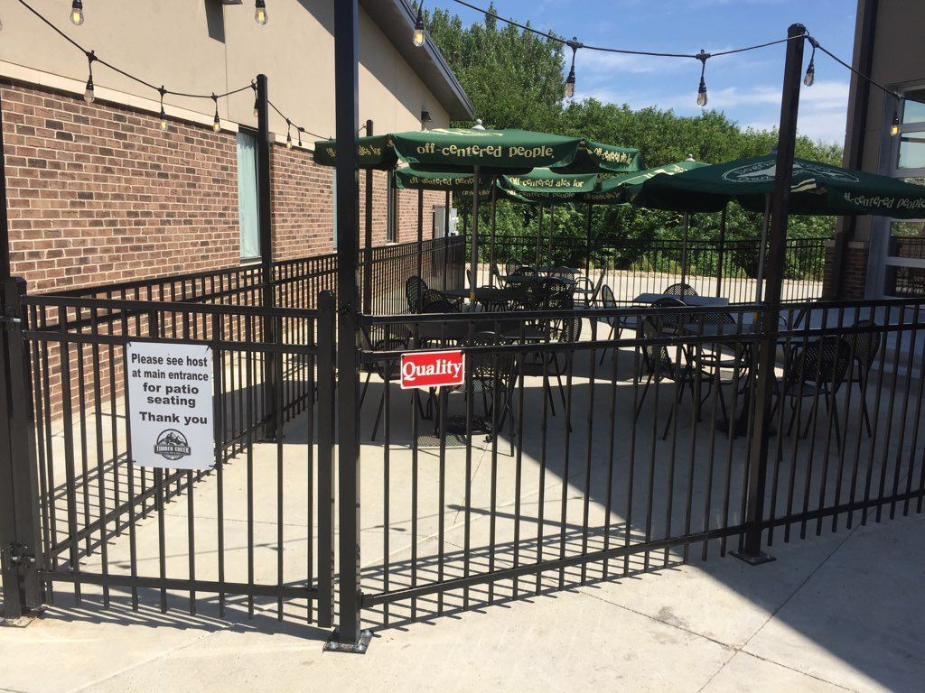 Outdoor patio with tables, chairs, and umbrellas enclosed by a black metal fence; a sign reads 