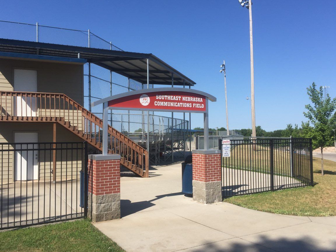 Entrance to a baseball field, featuring a covered sign, brick pillars, and a black fence under a clear sky.