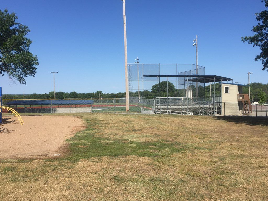 Baseball field with chain-link fence, dugout, and bleachers on a sunny day. A playground is in the foreground.