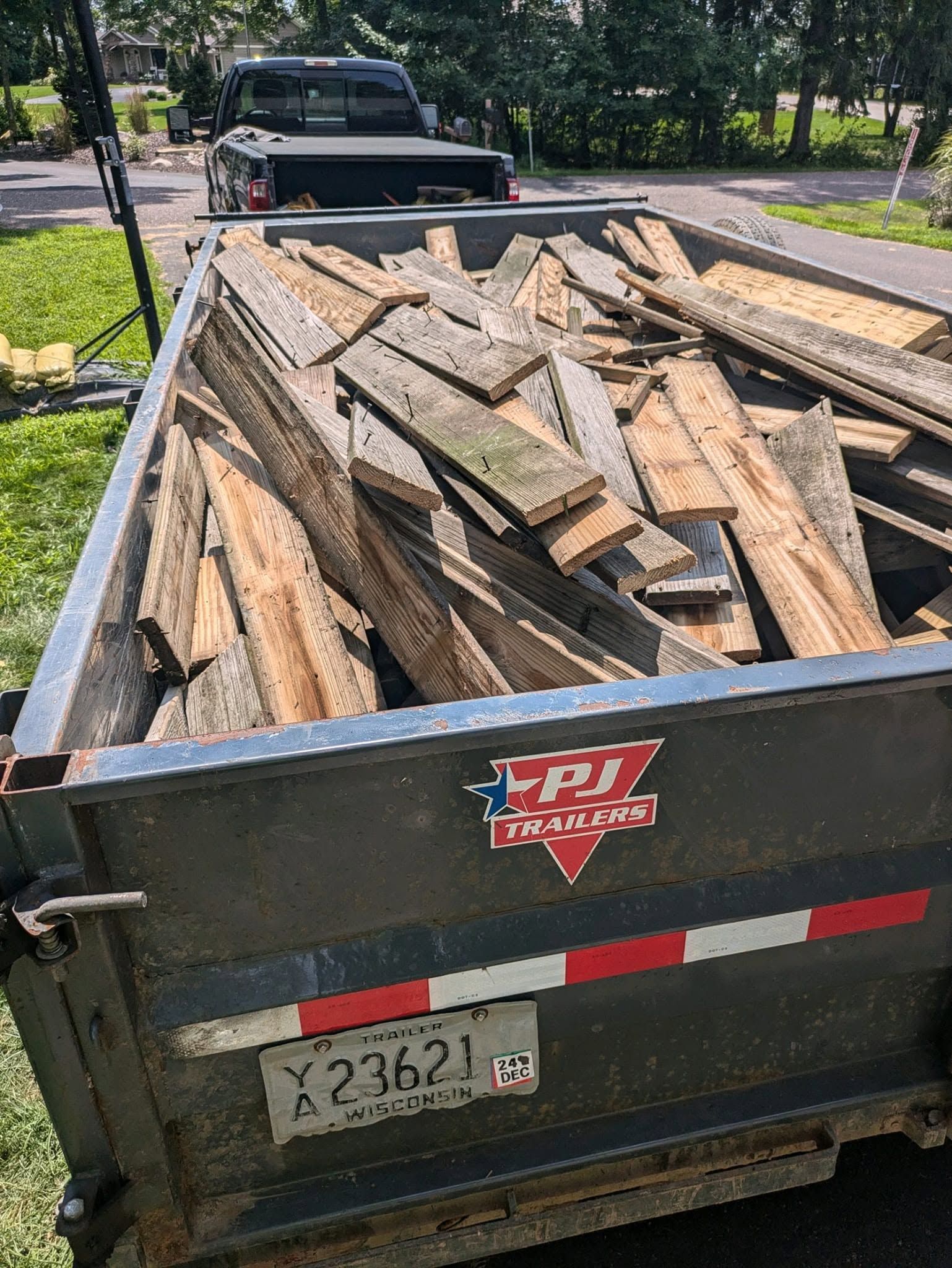 Dump trailer filled with old wooden planks on a driveway near parked trucks