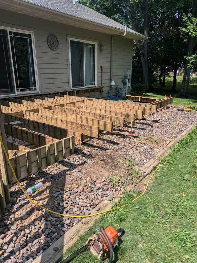 Wooden deck framing under construction beside a house, with tools and gravel along the lawn.