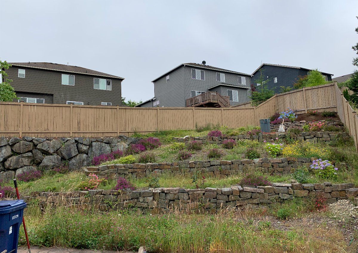 A wooden fence surrounds a lush green hillside with a blue trash can in the foreground.