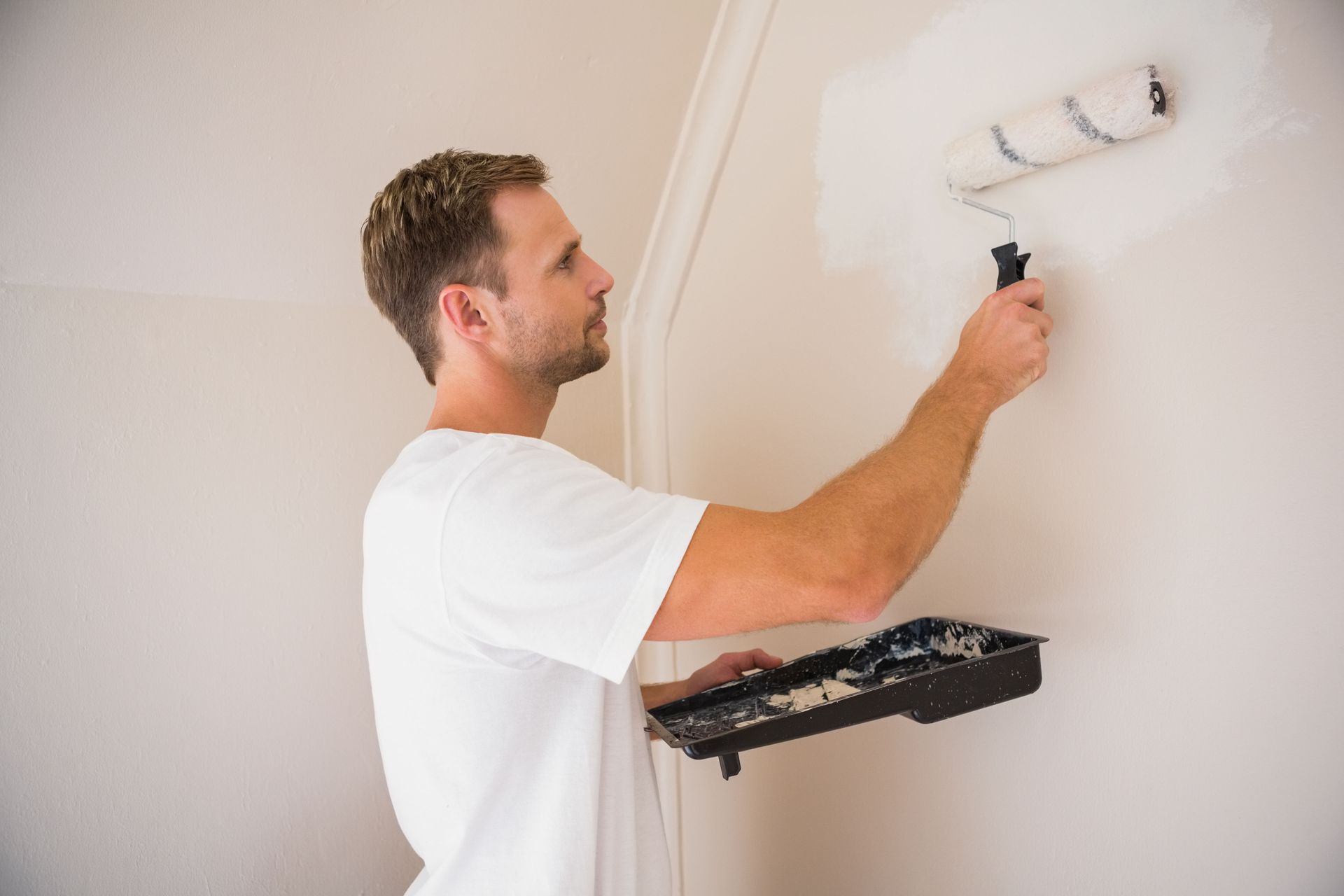 Man painting a white wall with a paint roller in a room.