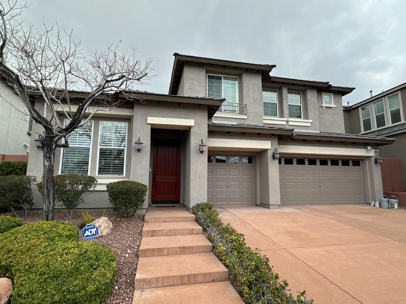 Two-story house with tan stucco exterior, red door, and brown garage doors. Brick steps lead to the entrance.