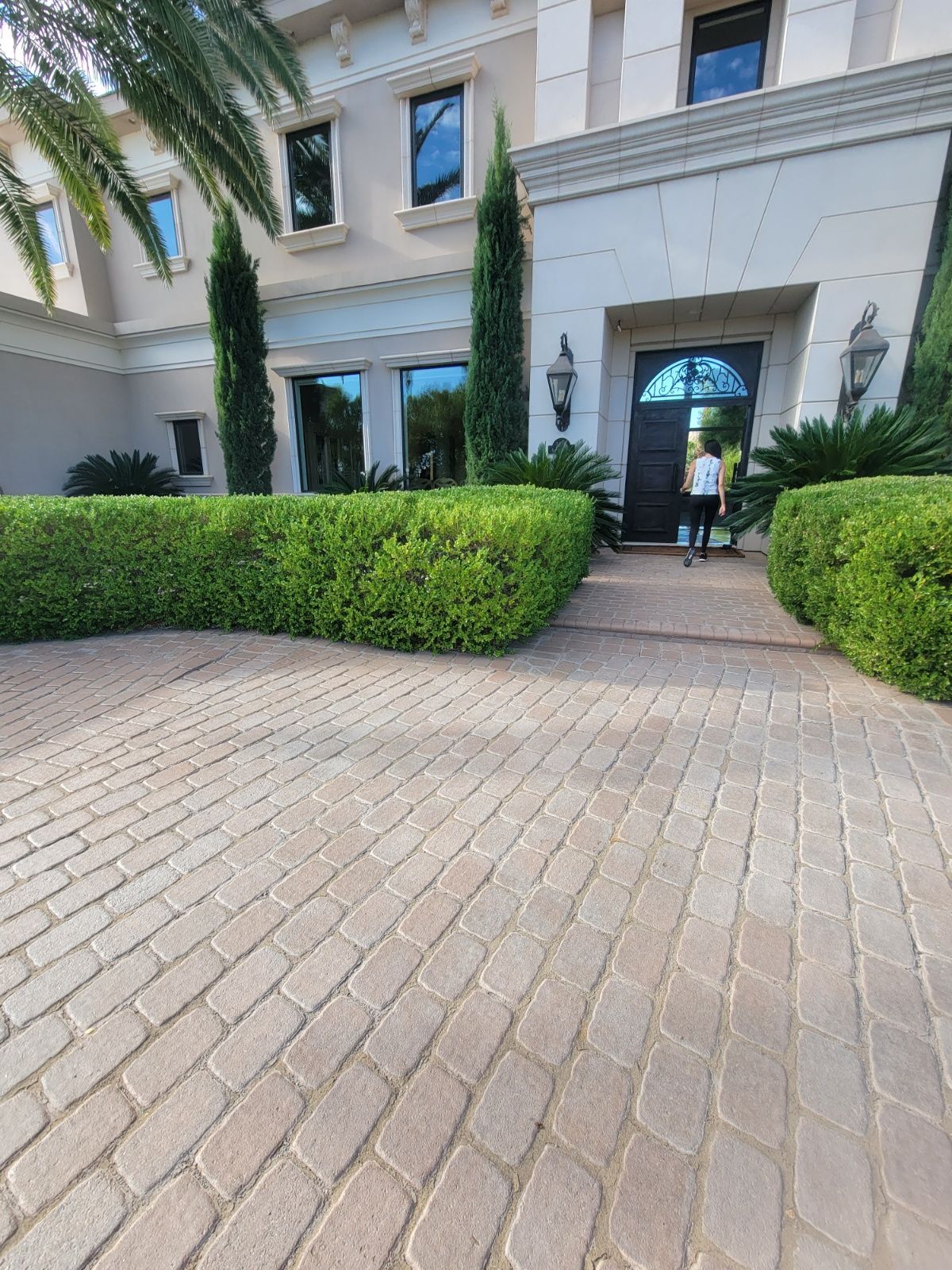 Brick driveway leading to a house with tall hedges and a person standing by the door.