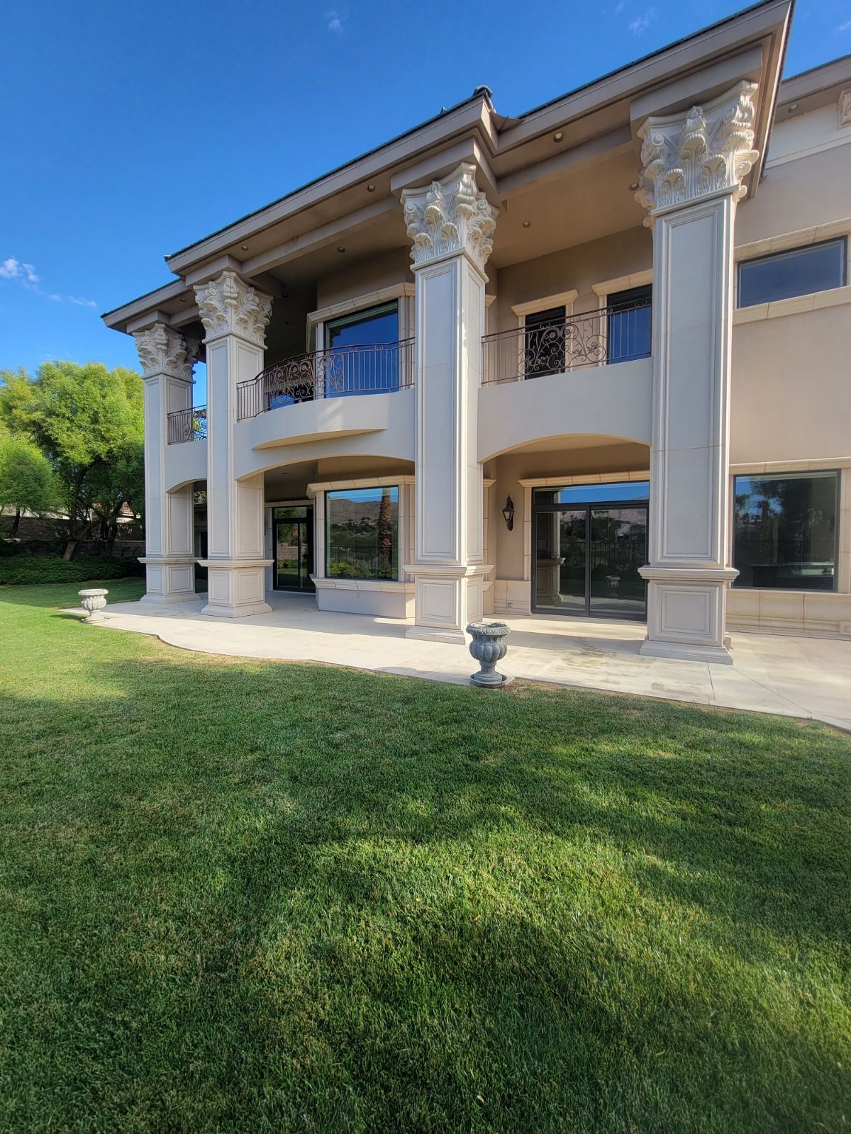 Two-story beige house with large columns. Green lawn in foreground, blue sky above.