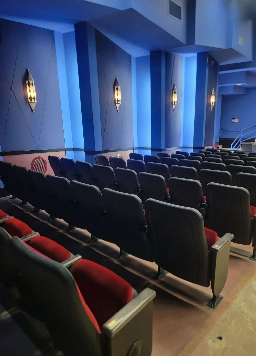 Rows of theater seats facing a stage with wall sconces; blue walls, red seats.