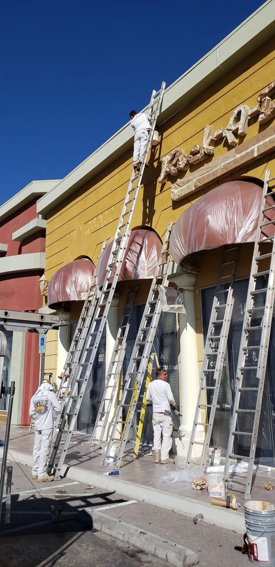Painters on ladders, painting the exterior of a commercial building a yellow color. Clear blue sky overhead.