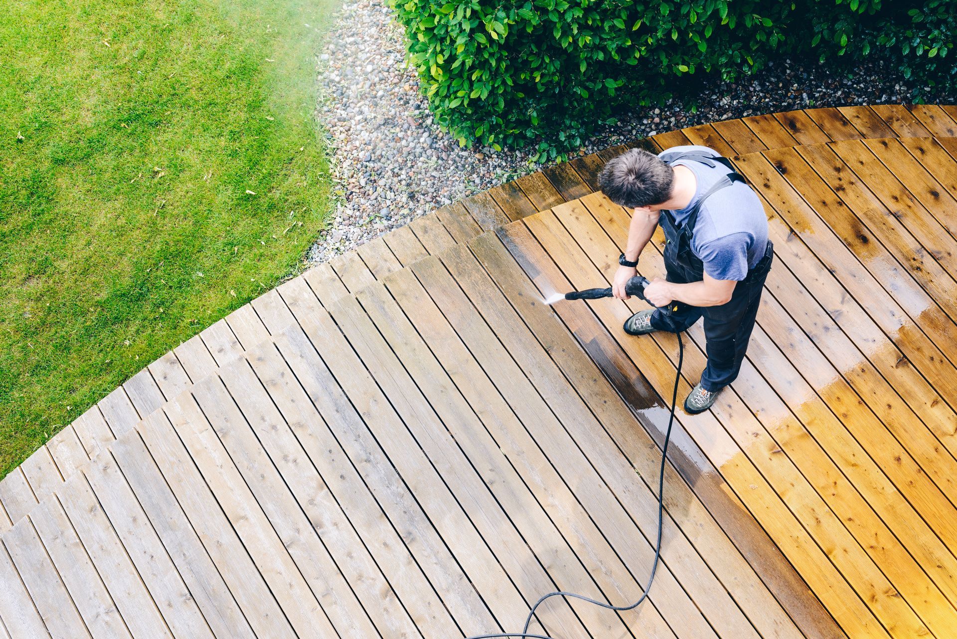 Person power washing a wooden deck, with water spray. One side is clean. Outdoors, green grass and plants.