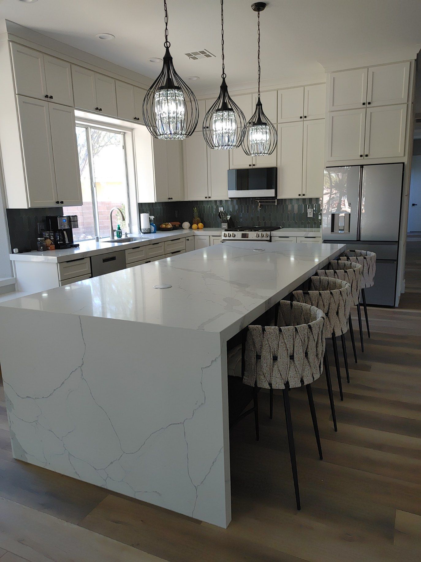 Modern kitchen with large white island, pendant lights, and bar stools. Light wood floors and stainless steel appliances.