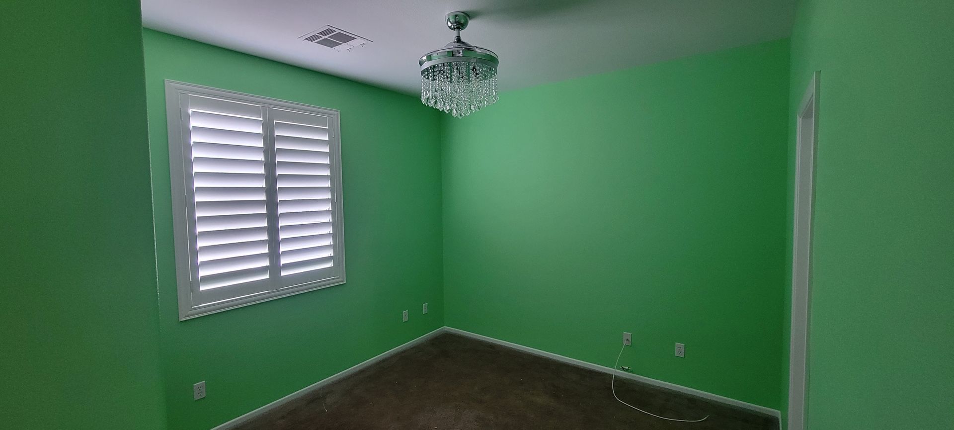 An empty room with green walls, a window with white shutters, and a crystal chandelier.