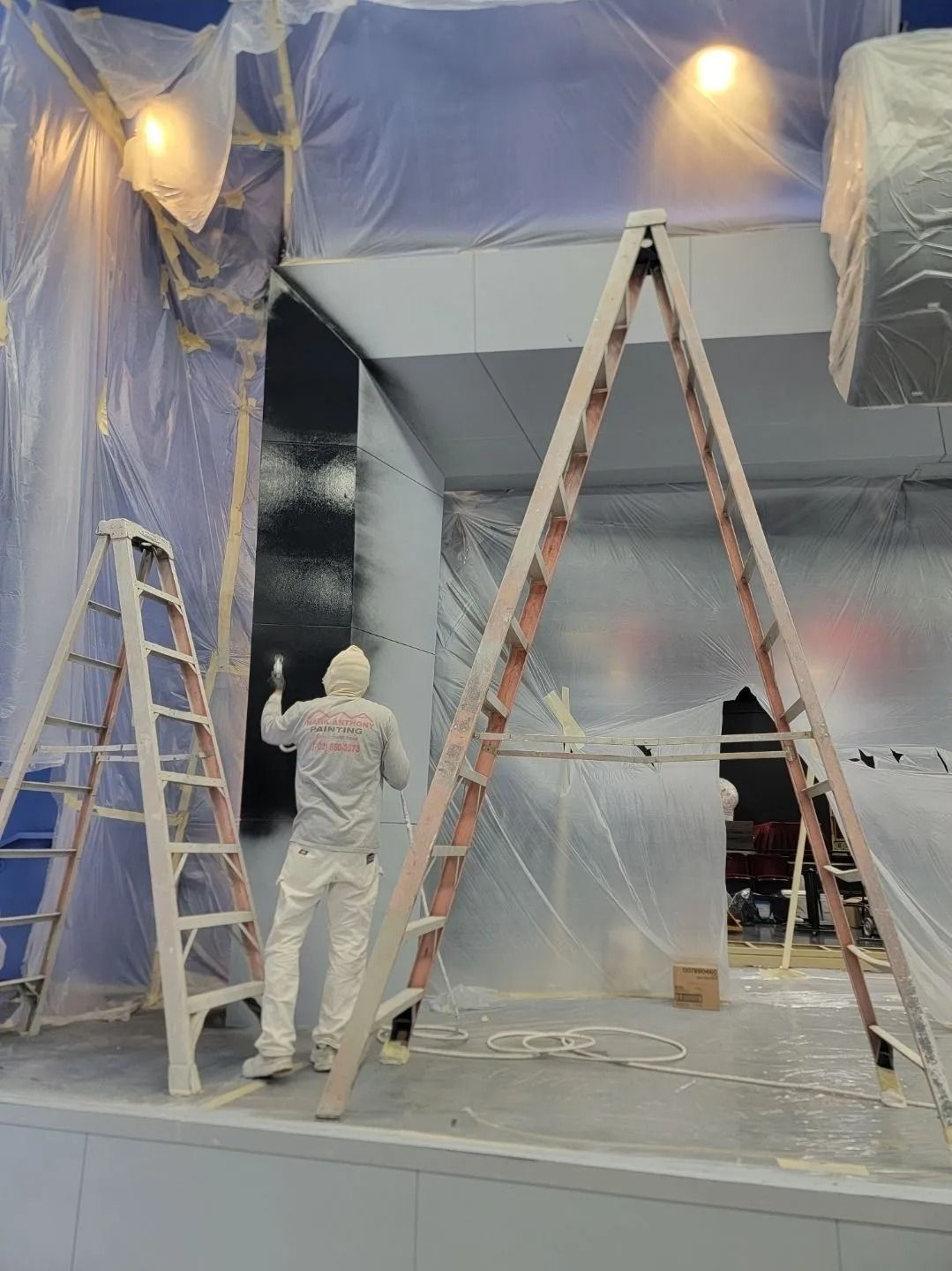 Person in white coveralls spraying paint, plastic sheeting covering a room, two ladders.