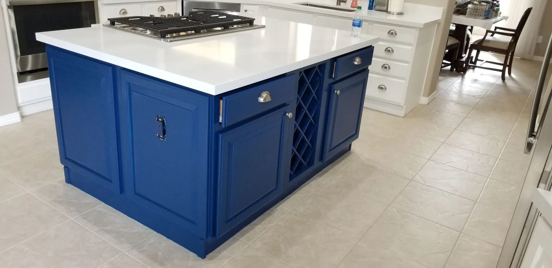 Blue kitchen island with white countertop, cooktop, and wine rack. Beige tiled floor.