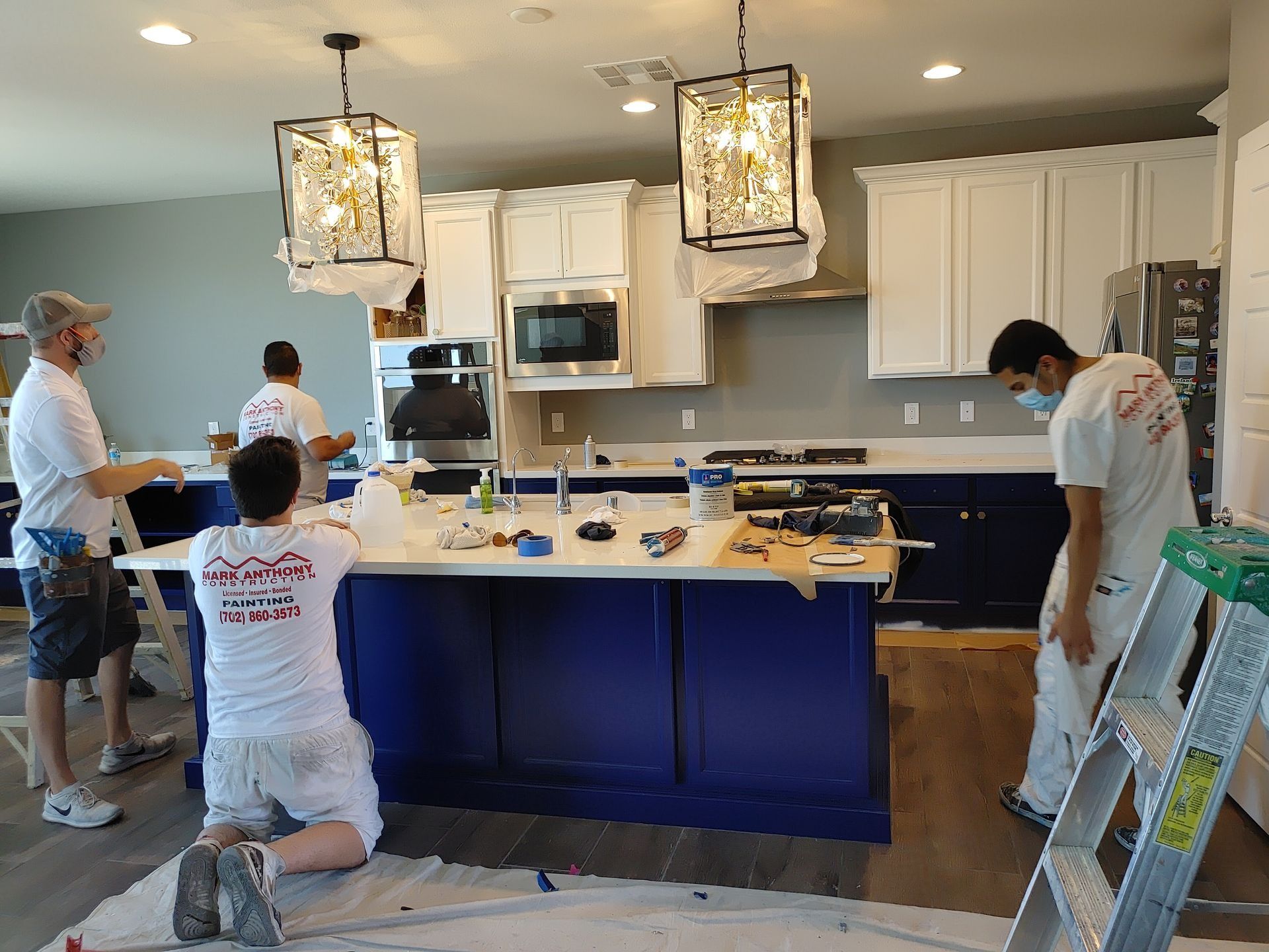 Four people painting a kitchen with blue and white cabinets; island and walls are being painted.