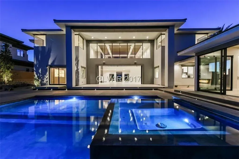 Modern home with pool at dusk, large windows, and a jacuzzi. Blue water reflects house lights.