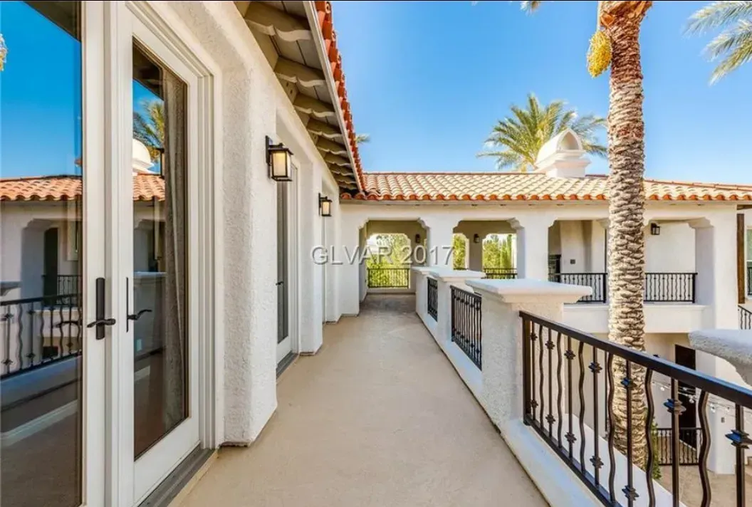 Balcony with white walls, iron railing, and tiled roof under a blue sky, palm trees nearby.
