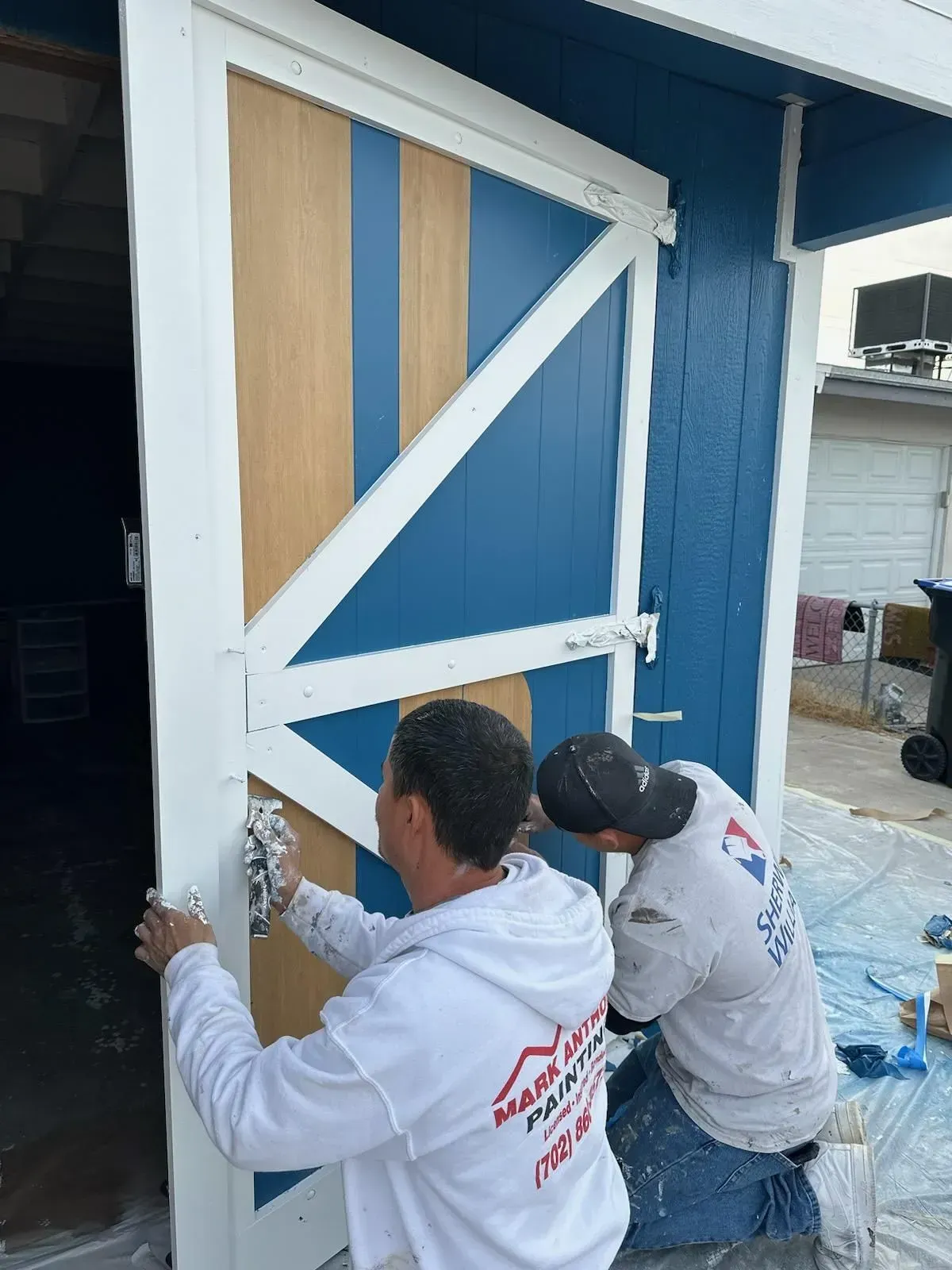 Two painters spray paint a blue and wood-tone barn door with white trim, outdoors.