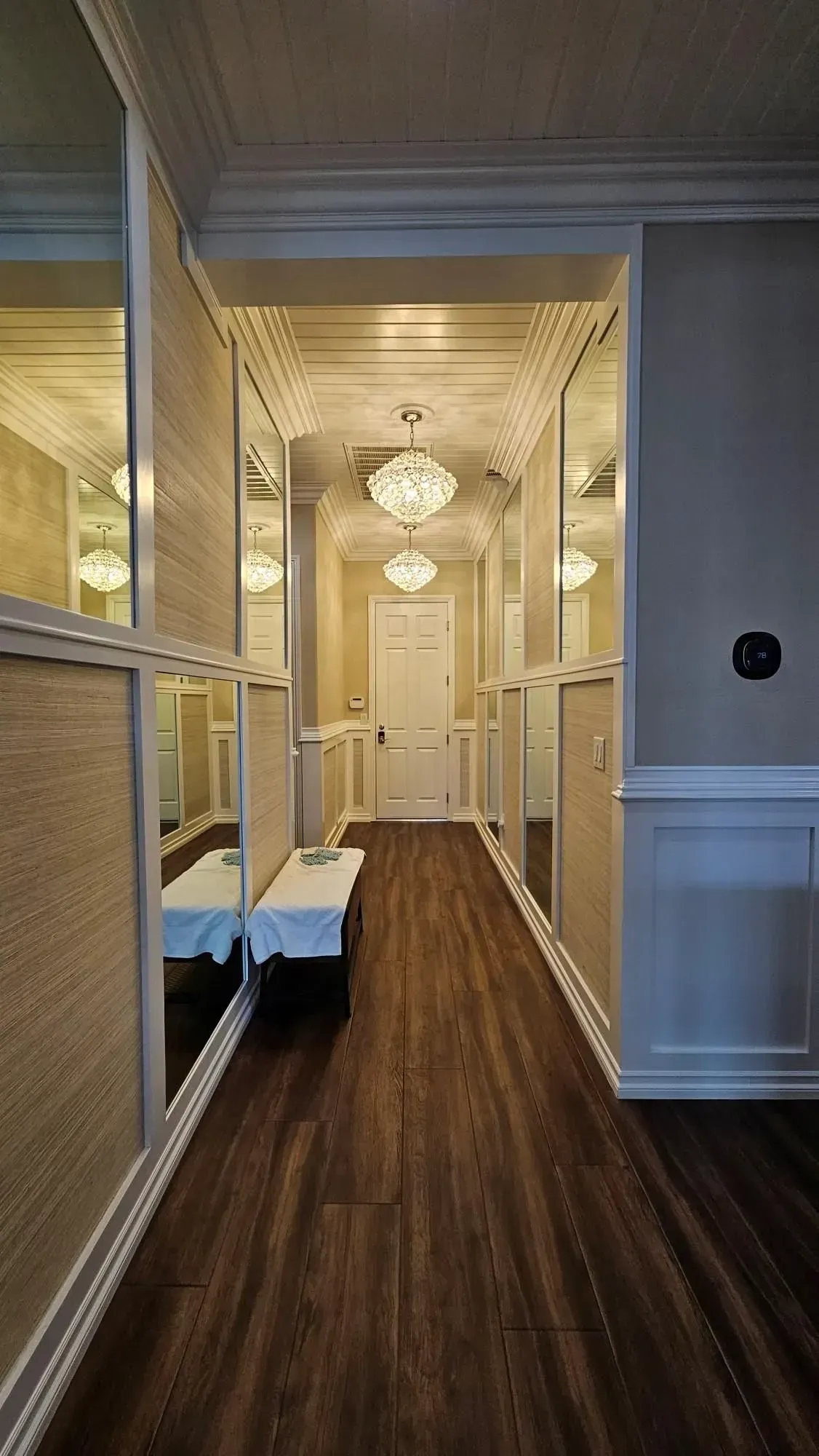 Long, mirrored hallway with a bench, chandeliers, and wooden floor. White door at the end.