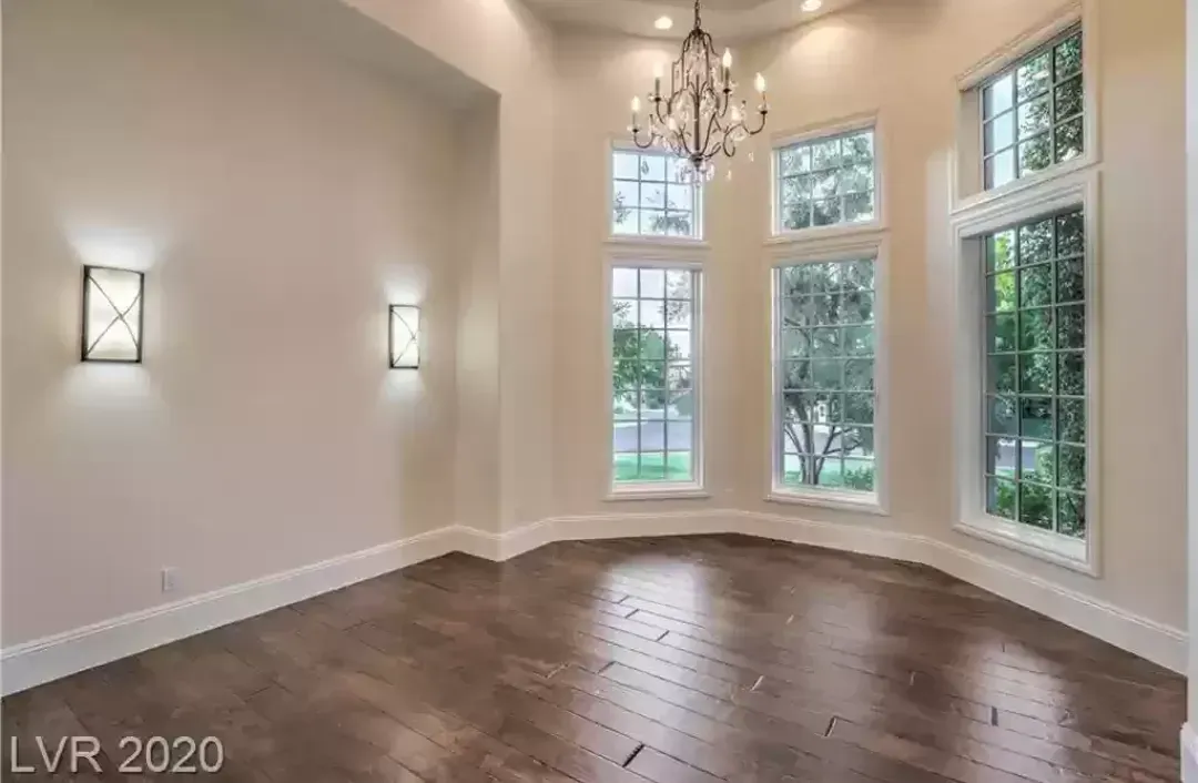 Empty room with hardwood floors, white walls, large windows, chandelier, and wall sconces.