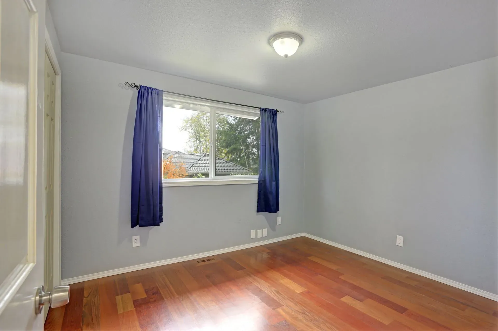 Empty room with gray walls, hardwood floor, and a window with blue curtains.