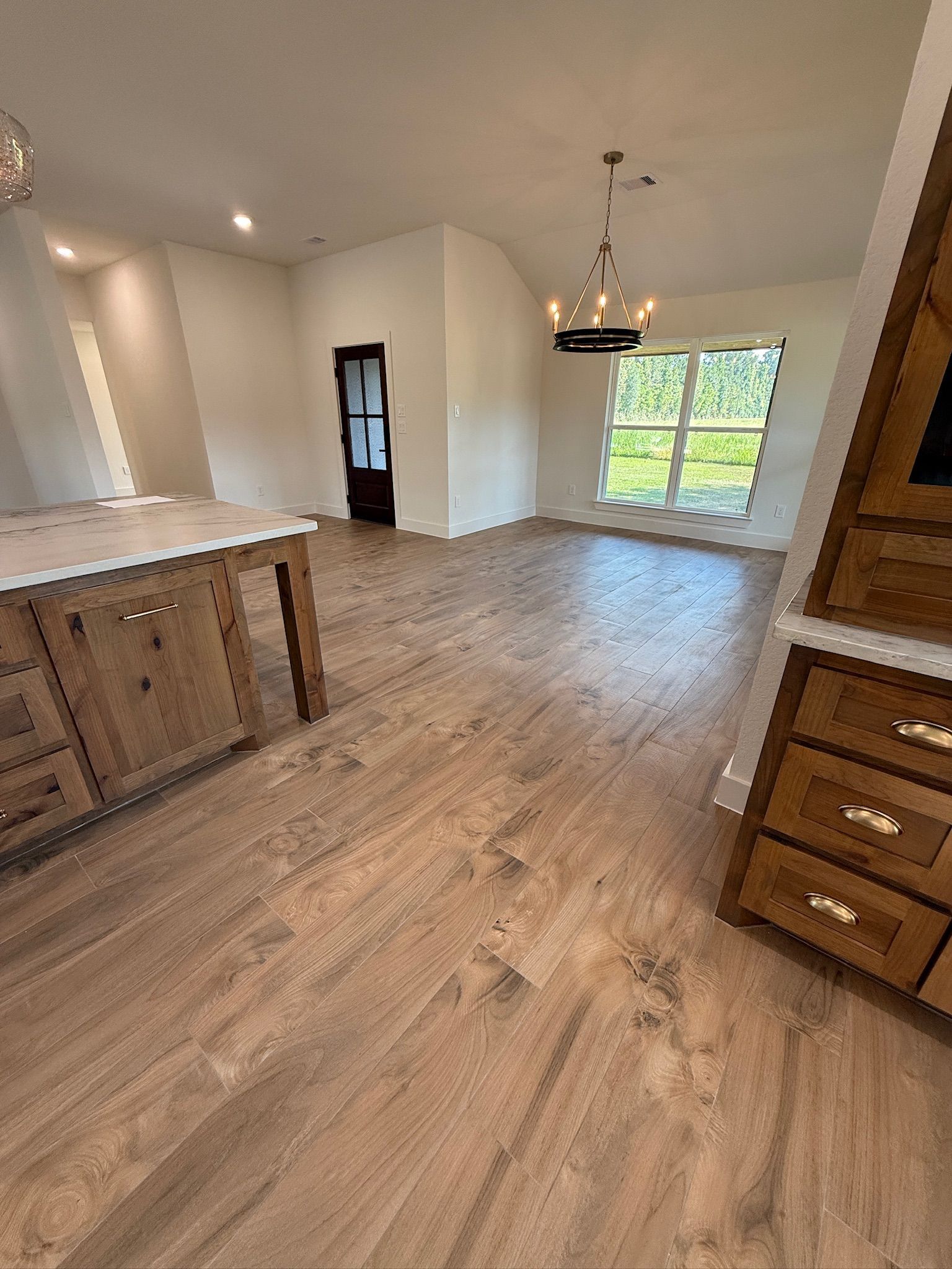 Empty modern kitchen and dining area with wood floors, island, chandelier, and large window.