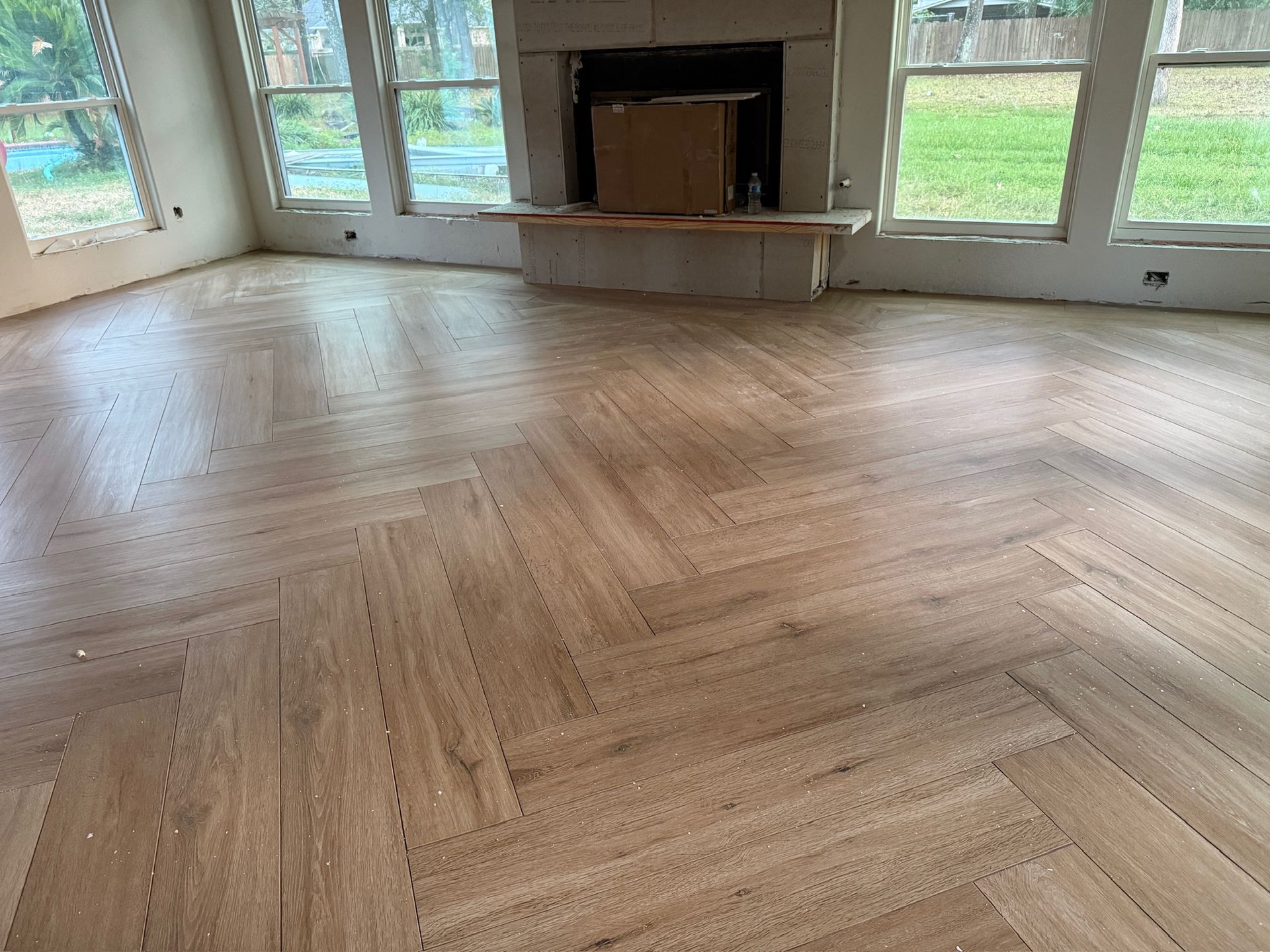 Empty living room with herringbone wood floor, large windows, and a central fireplace