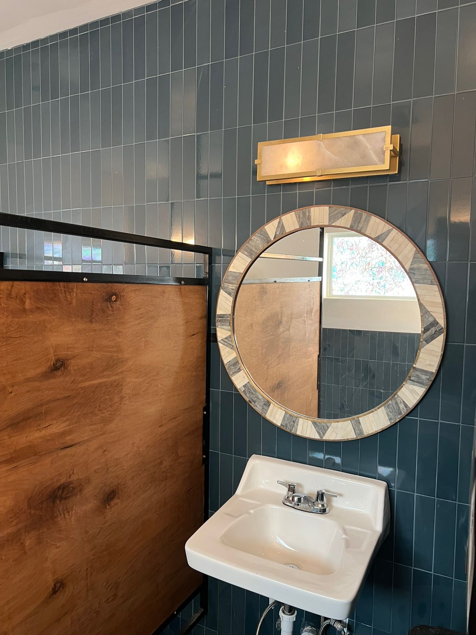 Small bathroom sink with round mirror, blue tile wall, wood partition, and brass light fixture