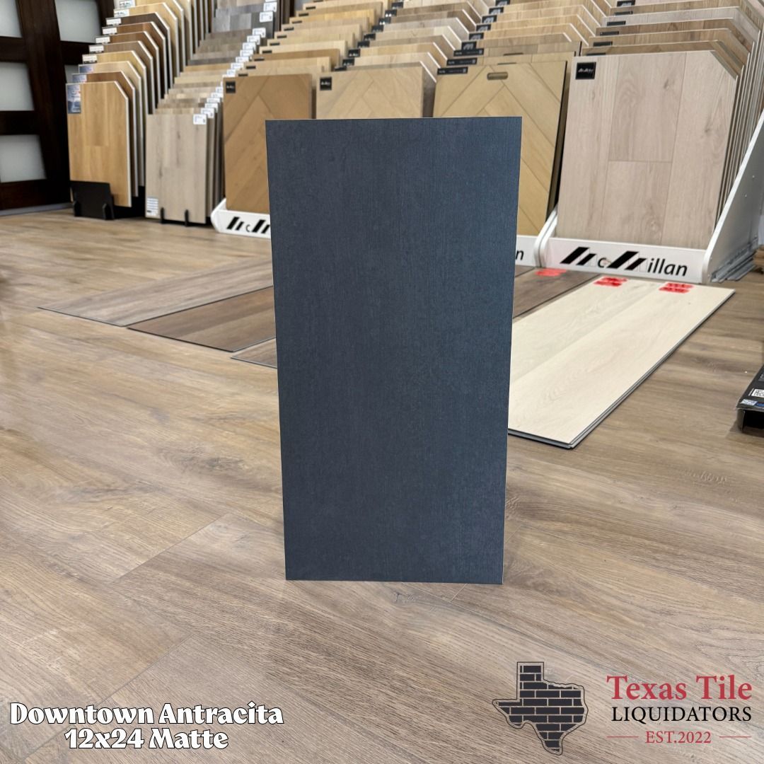 Dark gray rectangular tile sample standing upright in a tile showroom with wood-look flooring displays in the background