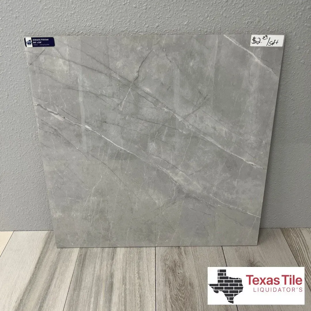A square grey tile with white marble-like veining, displayed on a wooden-look floor in front of a grey textured wall.