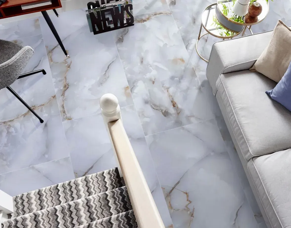 High-angle view of a living room with marble-patterned flooring, a light gray sofa, and a staircase with a zigzag runner.