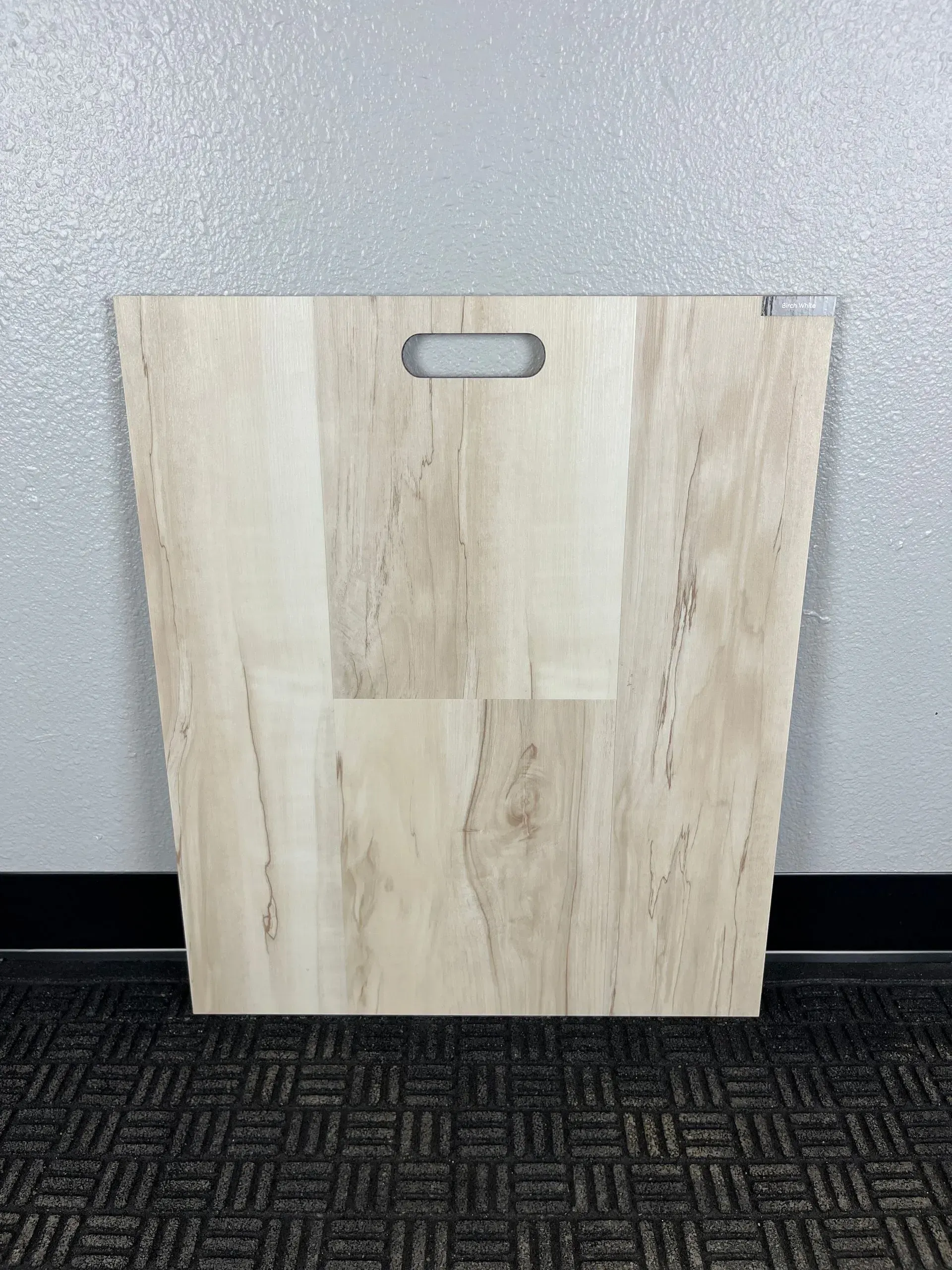 A square light wood-grain flooring sample board with a cut-out handle at the top, standing against a wall.