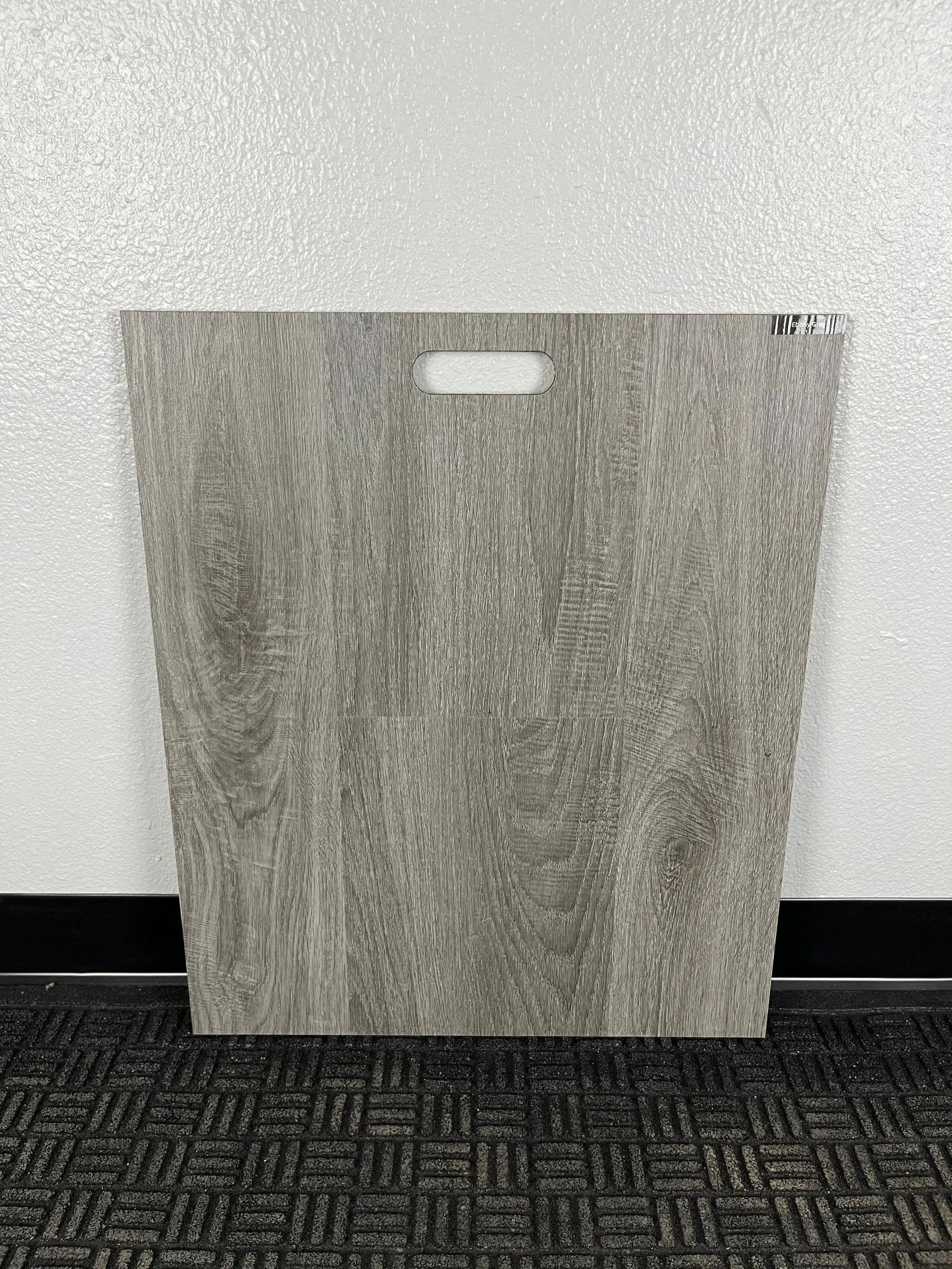 A rectangular, wood-grain sample board with a cutout handle, standing against a wall on a dark carpeted floor.
