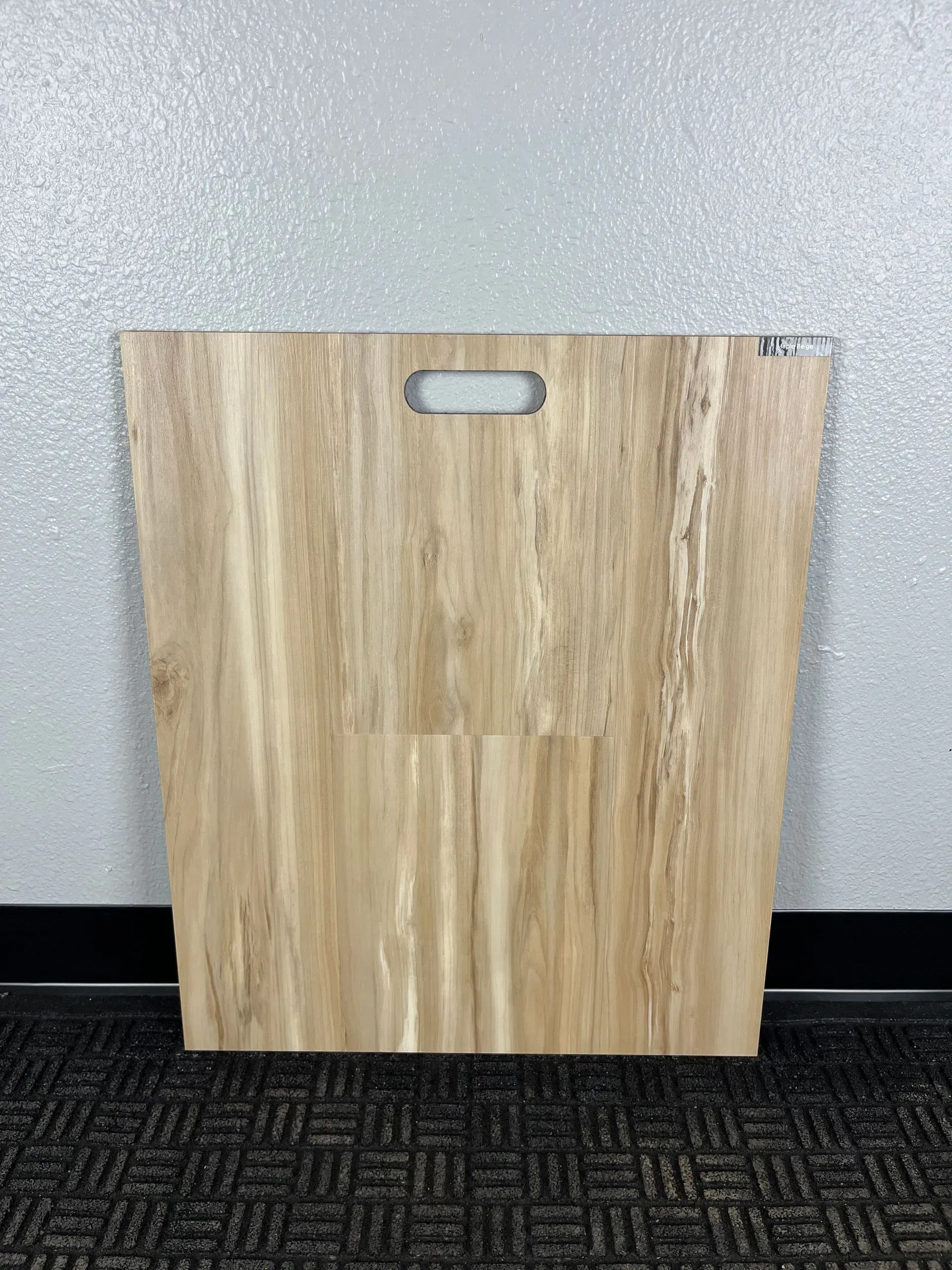 A light wood-grain flooring sample board with a handle cutout, placed against a textured gray wall and black carpet.