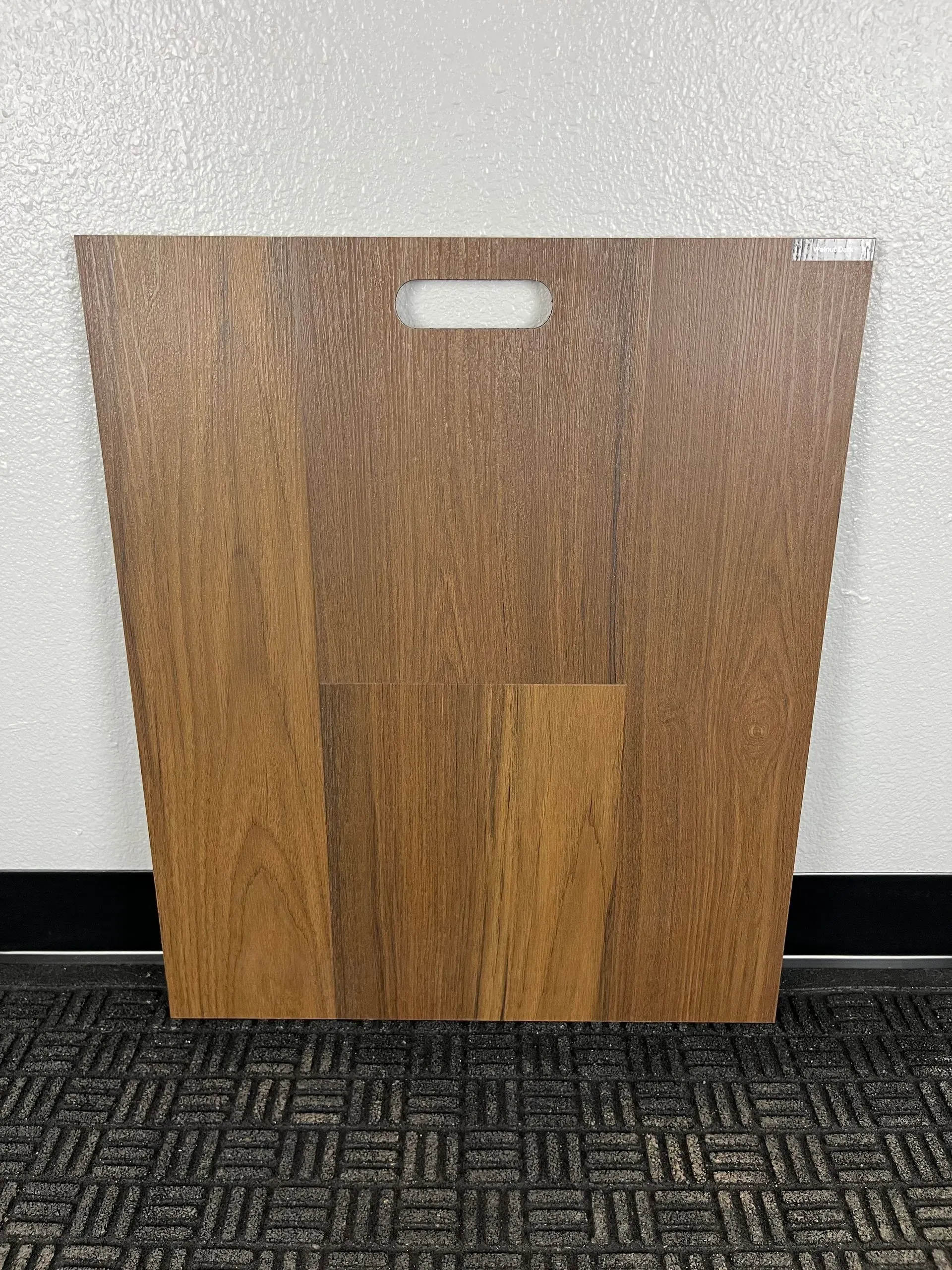 A rectangular wood-grain flooring sample board with a cutout handle at the top, placed against a wall and textured mat.