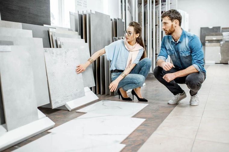 A woman and man in a tile showroom viewing and discussing gray ceramic flooring samples displayed on racks and the floor.