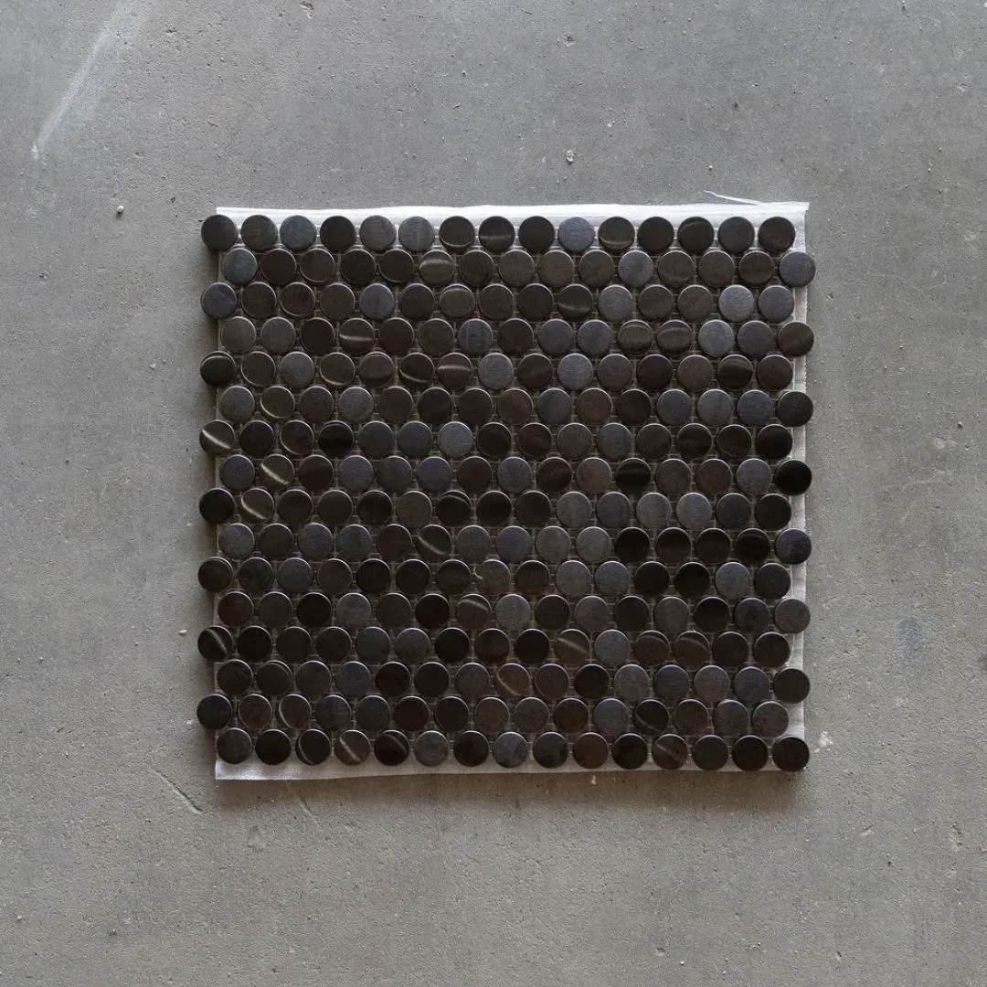 A square mesh sheet of small, circular, dark brown mosaic tiles placed on a gray concrete surface.